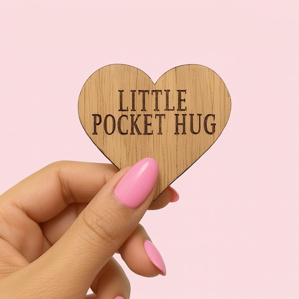 A hand holding a wooden heart-shaped object with the text "LITTLE POCKET HUG" engraved on it.