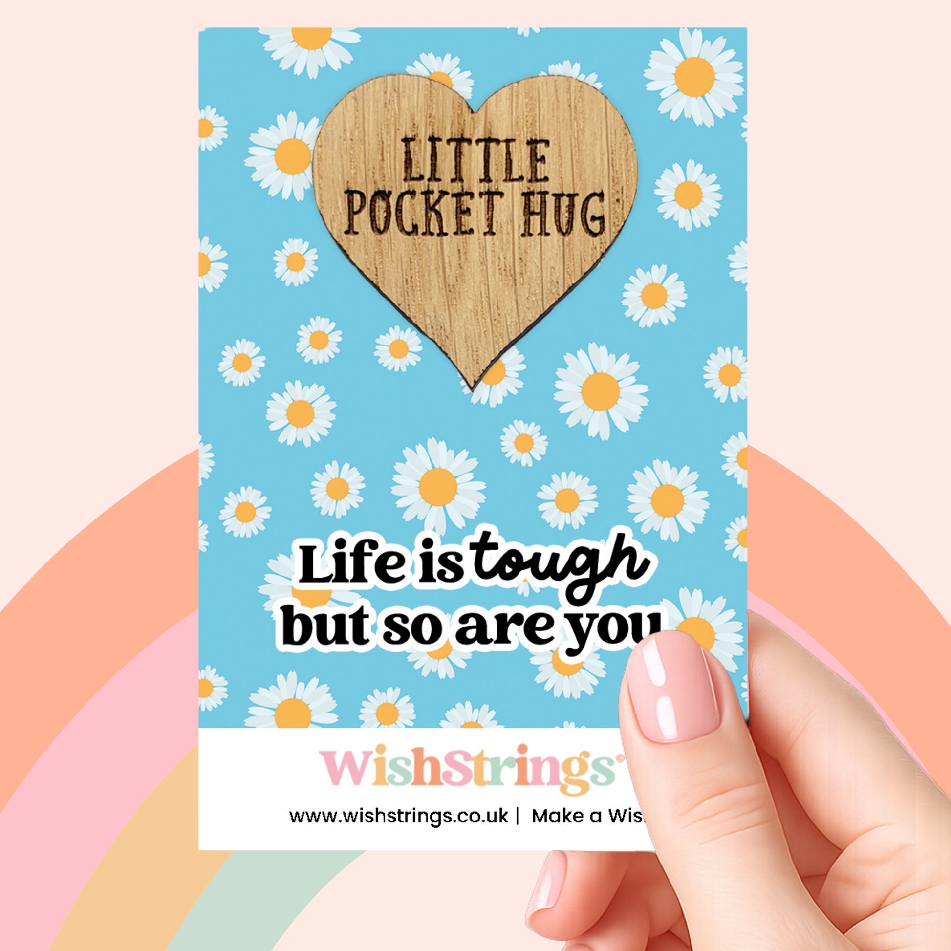 A hand holding a card with a heart-shaped wooden charm that says "Little Pocket Hug" and a message about life being tough but so are you.